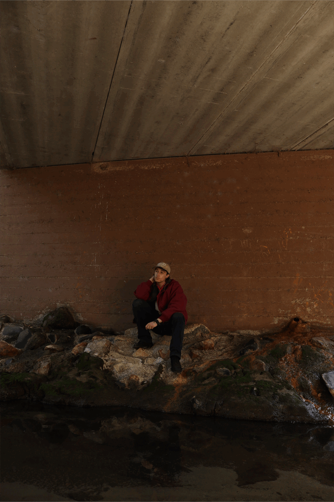 A person sits on rocky terrain beneath a concrete overpass